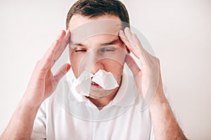 Young sick man isolated over white background. Portrait of guy with tissues in nose holding hands on headache. Guy