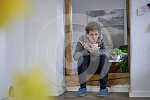 Young sick man with thermometer, blowing into his tea while sitting in a door frame, wearing a scarf