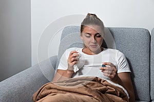 Young sick stressed woman lying at home covered with blanket checking her body temperature. Ill female have common cold and flue m