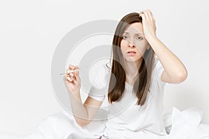 Young sick tired sad woman in pajamas sitting on bed, holding clinical thermometer with high fever temperature isolated