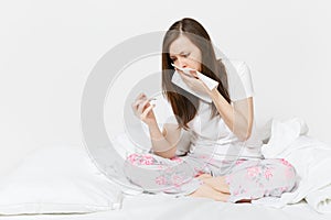 Young sick tired sad woman in pajamas sitting on bed, holding clinical thermometer with high fever temperature isolated