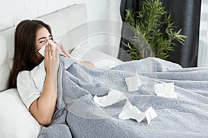 Young sick woman sitting in bed bowing out her running nose with paper towel feeling bad early in the morning