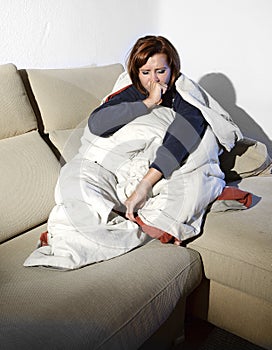 Young sick woman sitting on couch wrapped in duvet and blanket feeling miserable