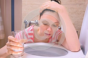 Young sick woman is vomiting in toilet sitting on the floor at homewith water glass in hand, food poisoning symptom.