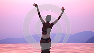 Young silhouette woman sitting in infinity swimming pool and waving two hands . she enjoying alone sunset
