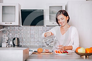 Young sincerely smiling female pouring a purified mineral water into transparent glass decanter at modern kitchen. Plenty of