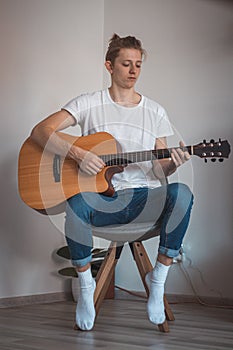 Young singer sits on a small stool in the corner and strums his song. Candid portrait of a blond man of European descent playing a