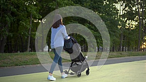 A young single mother walks with a baby in a stroller in the park. A woman is pushing a stroller with her son. View from