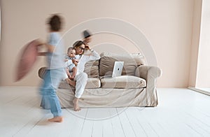 Young single mother working on laptop in loft sitting on couch while her sons running around her and shouting.