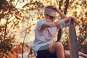 Young skater boy in sunglasses dressed in t-shirt and shorts sitting on the stone guardrail outdoors at a sunset.