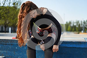 Young skater female putting on protective knee pads in a skatepark. Beautiful red haired person preparing for a ride in a outdoor