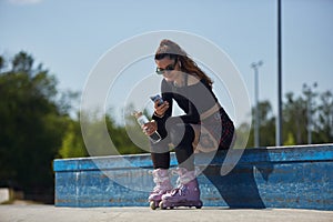 Young skater female sitting on a ledge in a skatepark with a glass bottle of water and smartphone. Roller blader person wearing