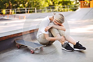 Young skater girl holding her painful leg and crying with skateboard near at skatepark isolated