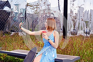 Young skater woman in blue dress making selfie by smartphone having coffee in a paper cup, sitting outdoor in a park.