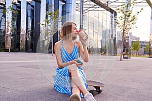 Young skater woman in blue dress with open shoulders having coffee in a paper cup, sitting outdoor in a park.