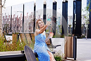 Young skater woman in blue dressmaking selfie by smartphone having coffee in a paper cup, sitting outdoor in a park.