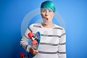 Young skater woman with blue fashion hair holding skateboard over blue isolated background scared in shock with a surprise face,