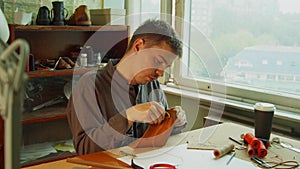 A young skilled tailor sews the element to a leather bag using a tailor`s needle. In the interior of a leather workshop