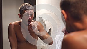 A young sleepy man brushes his teeth in the bathroom in front of a mirror