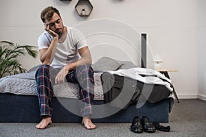 Young sleepy man in home clothes sitting on bed after waking up