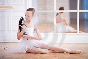 Young slender ballerina in white tutu sitting on floor with tiny Chihuahua in her hands in beautiful white room in front of mirror