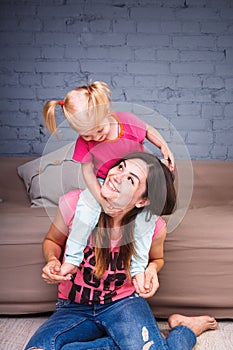 A young slender beautiful mother with her blond daughter on her shoulders play, enjoy and indulge in the teams near the sofa on th