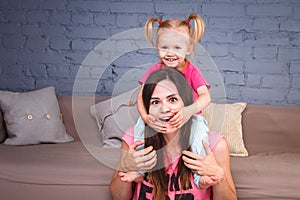 A young slender beautiful mother with her blond daughter on her shoulders play, enjoy and indulge in the teams near the sofa on th