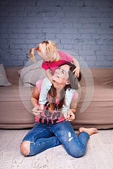 A young slender beautiful mother with her blond daughter on her shoulders play, enjoy and indulge in the teams near the sofa on th