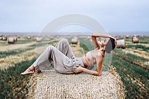 Young slender brunette girl with bright makeup in beige top with bare stomach and gray pants, lying on hay, posing for camera,