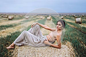 Young slender brunette girl with bright makeup in beige top with bare stomach and gray pants, lying on hay, posing for camera,