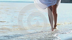 Young slender girl with bare feet walking model gait on sea sandy beach. Holding hands, white dress. surf hits woman on Shin and s