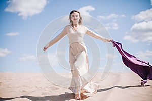 A young, slender girl in a beige dress with purple cloth in her hands posing in the desert in the wind