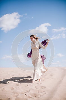 A young, slender girl in a beige dress with purple cloth in her hands posing in the desert in the wind