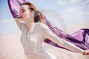 A young, slender girl in a beige dress with purple cloth in her hands posing in the desert in the wind