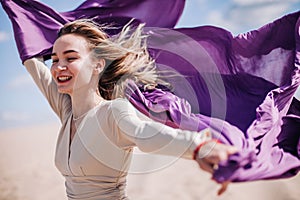 A young, slender girl in a beige dress with purple cloth in her hands posing in the desert in the wind