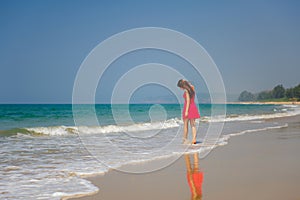 Young slender long-haired brunette in red dress walks barefoot on tropical beach along the surf against the sea and blue sky