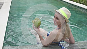 The young slender woman in Bikin drinks coconut milk from a coco on the edge of the pool in the tropical resort