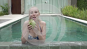 The young slender woman in Bikin drinks coconut milk from a coco on the edge of the pool in the tropical resort