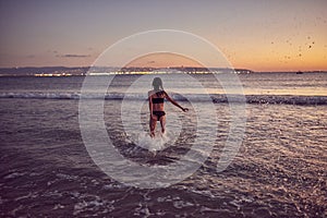 A young slender woman runs into the sea making splashes of water at sunset