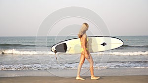 Young slender woman surfer in a bikini goes with a surfboard on the background of the sea