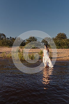 Young slender woman walks out of the beach going into the water holding a wet white summer dress exposing her legs to the knee
