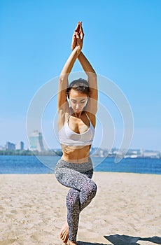 Young slim athletic woman makes yoga and balance exercises on the sand beach with city background. Healthy lifestyle