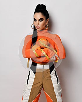 Young slim brunette woman in orange sportswear pants and turtleneck stands holding fresh oranges and measuring tape