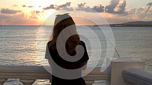 Young slim girl in a black t shirt standing by the sea leaning on railing during sunrise. Media. Rear view of a slim