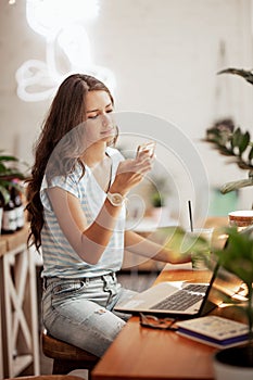 A young slim girl with long hair,wearing casual style, sits at the table with a laptop and looks at her phone in a cozy