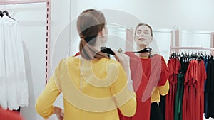 Young slim lady with ponytail choosing a dress in clothes shop. Woman shopping and checking clothes in front of mirror