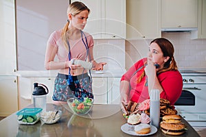 Young slim and overweight women look at each other in kitchen. Thin model hold bowl with salad in hands. Plus size woman