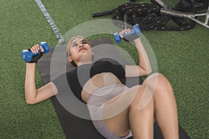 A young, slim and sexy asian woman does dumbbell floor presses while lying on the black mat. Working out and exercising upper body