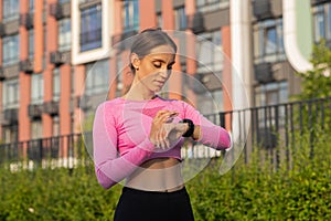 A young slim sportswoman stands on the city street, looks intently at her watch and prepares for a morning jog in a park