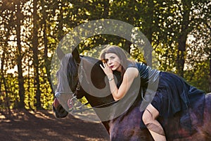 Young slim woman brunette in a black dress sitting on dark brown horse.